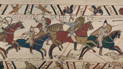 Section of the Bayeux Tapestry showing a group of warriors with shields and spears on horseback charging from left to right.
